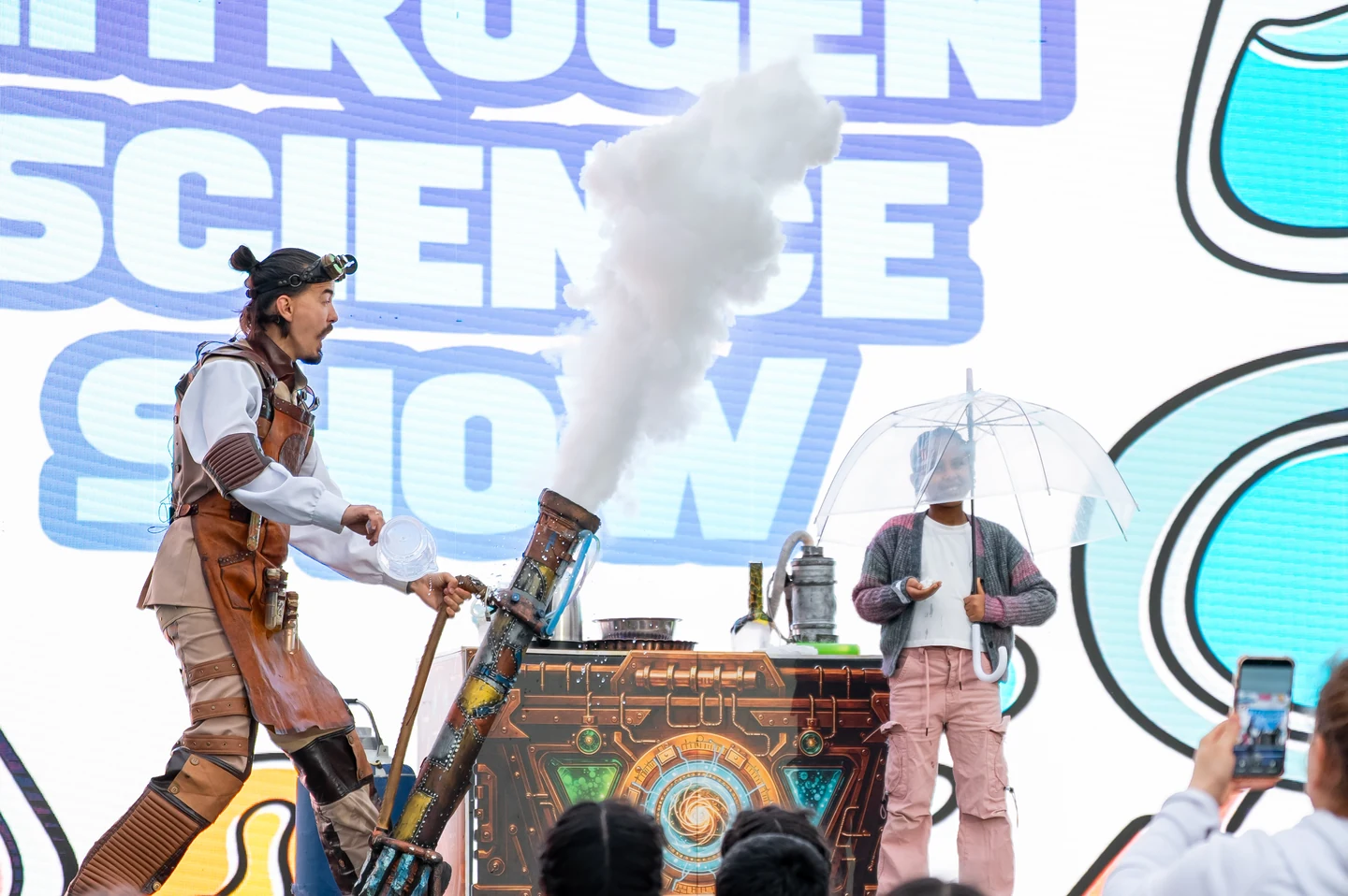 A child holding a clear umbrella joins the Science Show professor beside a towering cloud during a live experiment.